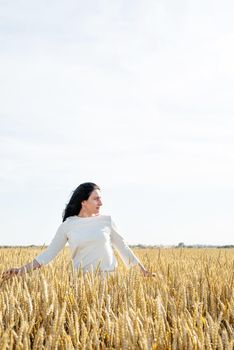 Mid adult woman in white dress walking and dancing across golden field holding heap of rye lit by sunset light, copy space