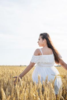 Young brunette woman walking across golden field holding heap of rye and wearing white dress lit by sunset light, copy space
