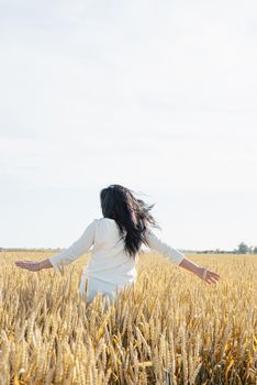 Mid adult woman in white dress walking and dancing across golden field holding heap of rye lit by sunset light, copy space