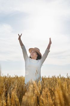 Mid adult woman in white dress walking and dancing across golden field holding heap of rye lit by sunset light, copy space