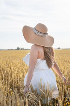 Young brunette woman walking across golden field holding heap of rye and wearing white dress lit by sunset light, copy space