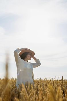 Mid adult woman in white dress walking and dancing across golden field holding heap of rye lit by sunset light, copy space