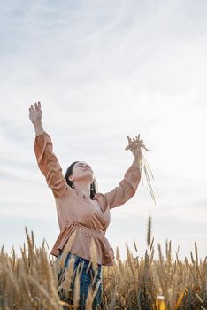 Mid adult woman in beige shirt walking across golden field holding heap of rye lit by sunset light, copy space