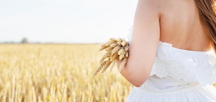 Young brunette woman walking across golden field holding heap of rye and wearing straw hat lit by sunset light, copy space