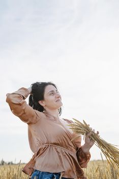 Mid adult woman in beige shirt walking across golden field holding heap of rye lit by sunset light, copy space