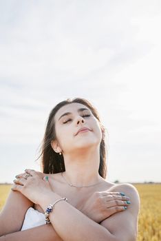 Young brunette woman walking across golden field holding heap of rye and wearing white dress lit by sunset light, copy space