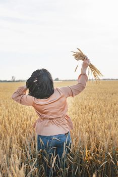 Mid adult woman in beige shirt walking across golden field holding heap of rye lit by sunset light, copy space