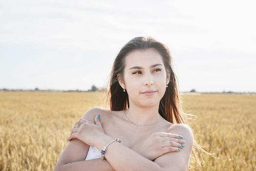 Young brunette woman walking across golden field holding heap of rye and wearing white dress lit by sunset light, copy space