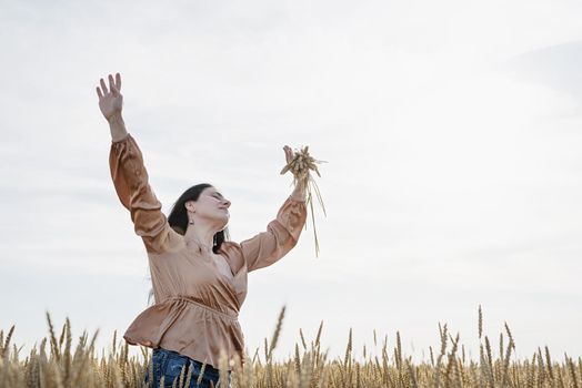 Mid adult woman in beige shirt walking across golden field holding heap of rye lit by sunset light, copy space