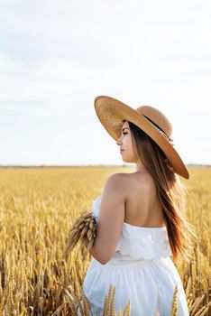 Young brunette woman walking across golden field holding heap of rye and wearing straw hat lit by sunset light, copy space