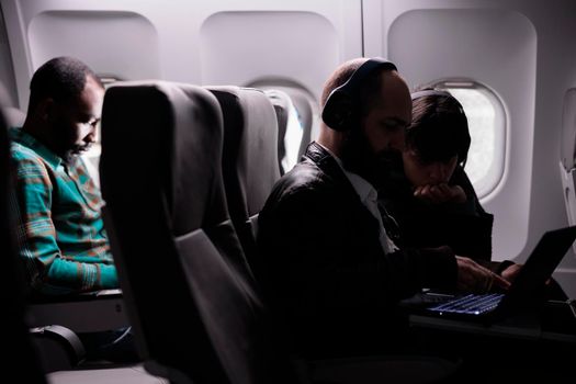 Diverse group of airplane passengers waiting to arrive at destination, flying in economy class. People using laptop and smartphone during international flight on plane, airline transport.