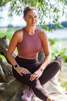 Sexy attractive woman in yoga pants and top sitting in forest taking rest after trainings, looking at camera with serious motivated concentrated expression. Relaxation after workout summer day outdoor