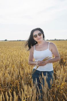 Mid adult brunette woman walking across golden field holding heap of rye lit by sunset light, copy space