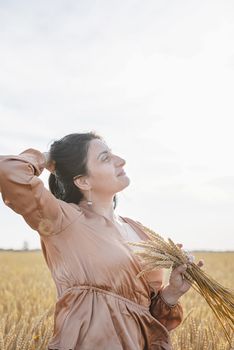 Mid adult woman in beige shirt walking across golden field holding heap of rye lit by sunset light, copy space