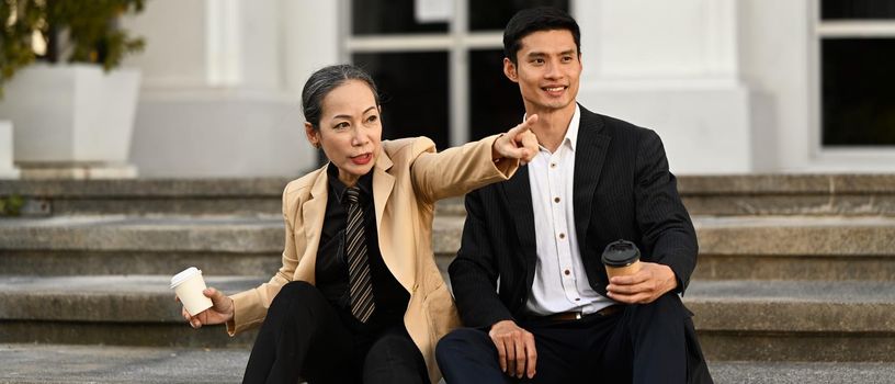 Shot of two mature businesswoman and businessman talking to each other, sitting on stairs of an office building in a beautiful day.