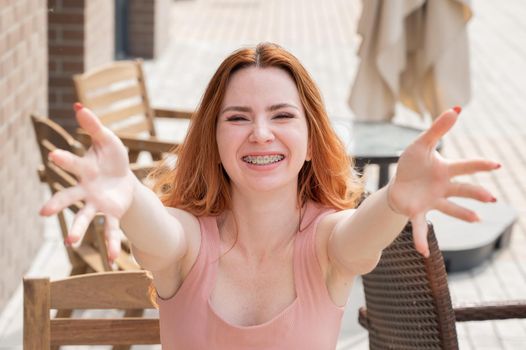 Young red-haired woman sits in a street cafe and stretches her arms to hug