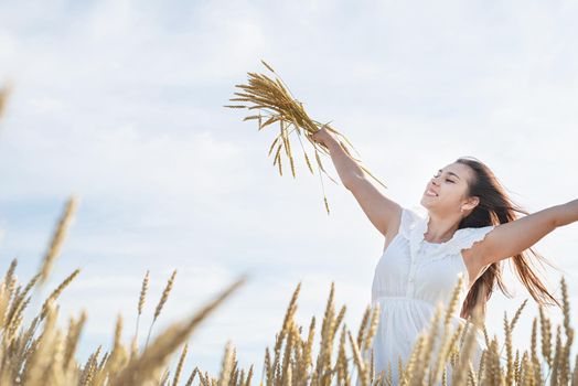 Young brunette woman standing in golden field holding heap of rye and wearing white dress lit by sunset light, laughing, copy space