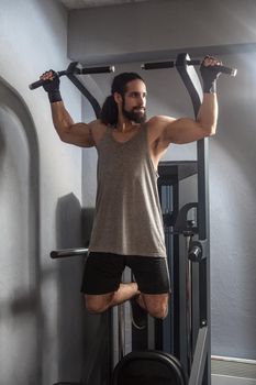 Full length portrait of confident young adult strong man with long curly hair working out in gym, hanging on horizontal bar with hands, doing pull ups with, exercises for triceps. indoor, looking away