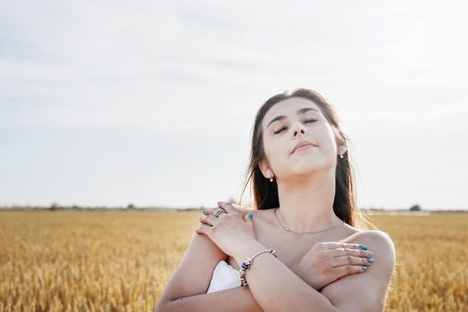 Young brunette woman walking across golden field holding heap of rye and wearing white dress lit by sunset light, copy space