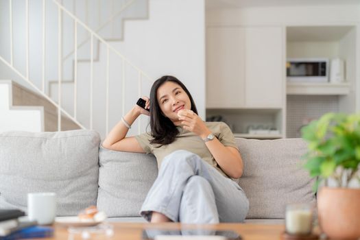 Image of a beautiful asian woman searching channel with remote control to watch tv while sitting on sofa at home.