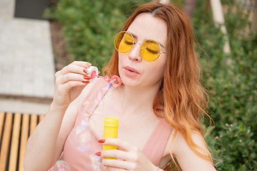 Young red-haired woman blowing soap bubbles outdoors. Girl in yellow sunglasses and braces