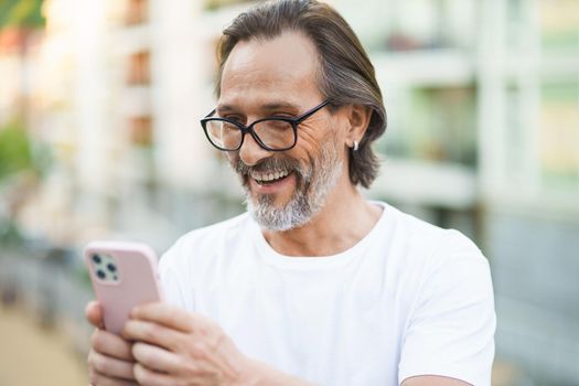 Mature man with grey beard video calling using smartphone camera standing outdoors with city streets background wearing white shirt. Happy middle aged man on street of european city with mobile phone.