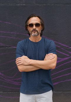 Portrait of handsome mature man standing next to dark urban wall with hands folded wearing sunglasses, dark blue t-shirt and light shorts. Mature business man or freelancer outdoor.