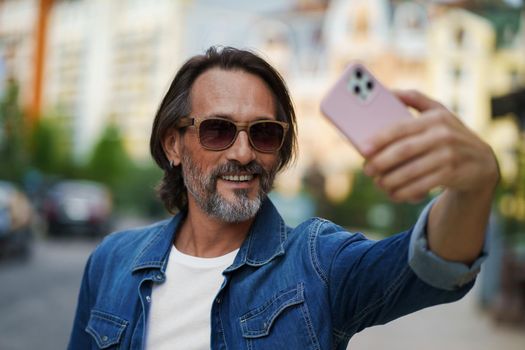 Middle aged handsome grey bearded metrosexual man smiling on camera old town wearing casual. Happy senior man on street of european city making selfie using mobile phone. Travel concept.