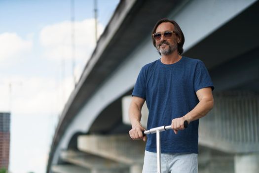 Handsome stylish mature man with grey beard stand on an town scooter having a ride on the streets under the bridge after work outdoors. Travel, lifestyle concept.