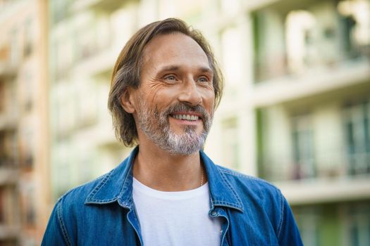 Cheerful handsome mature man with grey beard walking, touring urban, old town european streets wearing casual jeans shirt with white t-shirt. Travel lifestyle concept.