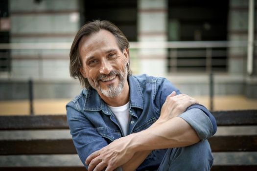 Happy smiling handsome grey beard middle age man sits at the bench with hands folded leaned on lifted knee outdoors at urban city background enjoying free time. Travel and people concept.