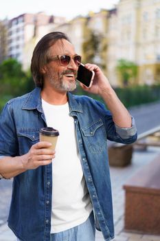 Excited mature man with grey beard talking on the phone while having coffee in paper disposable cup, work or travel time standing outdoors in urban city background wearing jeans shirt and sunglasses.