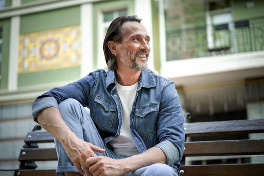 Handsome happy smiling middle aged man with grey beard sits on the bench with hands leaned on lifted knee looking away at old town background enjoying free time. Travel and people concept.