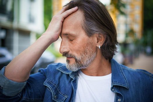 Handsome man overpass middle age crisis touching his forehead looking emotionally exhausted. Experiencing headache, stress mature handsome man standing outdoors european old town streets.