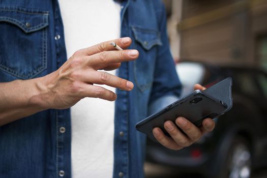 Smoking cigaret middle aged man checking text messages or social media using his smartphone wearing jeans shirt and white t-shirt standing outdoors on urban city background. No face visible.