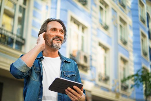 Mature man use wireless earphones talking or listening music while travel in old town streets. Handsome middle aged freelancer man using digital tablet having a call while walking on urban streets.