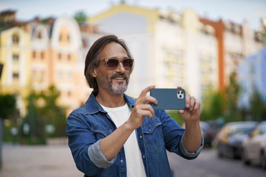 Happy middle aged grey bearded man taking pictures using smartphone camera in old town wearing casual. Happy senior man on street of european city with mobile phone. Travel concept.