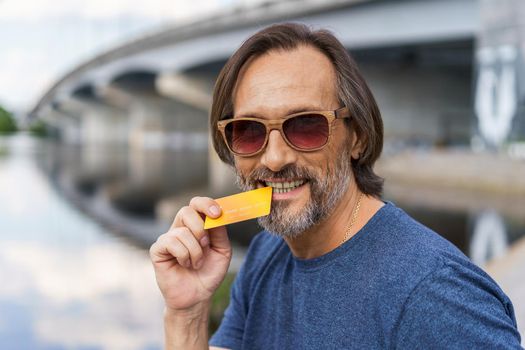 Handsome middle aged man standing outdoors at urban city background near the bridge over the river holding debit or credit card, shopping online or paying online banking. E-banking concept.