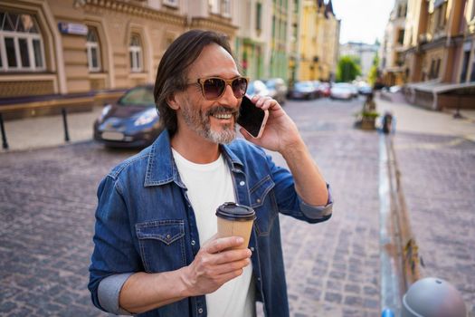 Happy mature grey bearded man talking on the phone, having coffee in paper cup or lunch break during work or travel time standing outdoors in urban old city background wearing jeans shirt.