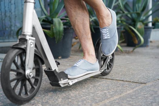 Male feet in a sneakers stand on an town adult scooter with big wheels having a ride on the streets or park after work outdoors with agave plants on background. No face visible. Focus on left leg.