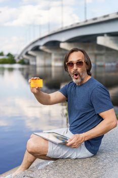 Excited freelancer man sitting outdoors next to river working using digital tablet pc and showing debit or credit card, shopping online or paying online banking. E-banking concept.