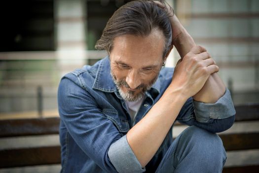 Mature grey bearded man deep in his thoughts with positive emotions on his face. Happy thinking of life middle aged man leaned his hands on knee looking down his head sitting on the bench in old town.