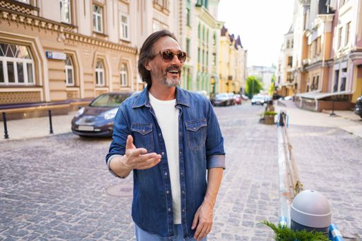 Portrait of a happy mature man outdoors in old town city smiling wearing jeans shirt and sunglasses. Happy mature man wearing sunglasses and looking sideways outdoor on a summer day.