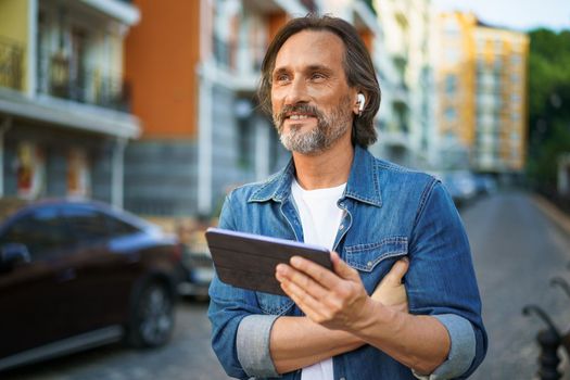 Handsome middle aged freelancer man using digital tablet having a call while walking on urban streets. Mature man use wireless earphones talking or listening music while travel in old town streets.