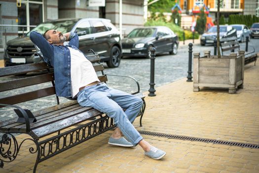 Relaxed handsome middle age man sits at the bench outdoors at urban city background enjoying free time with tablet standing next to him. Travel and people concept.