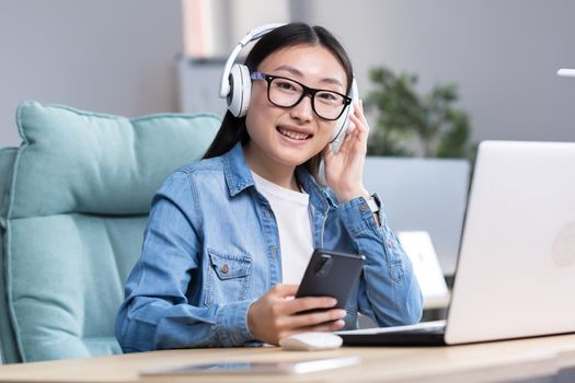 Portrait of young Asian female student studying online at laptop, using laptop and headphones to listen to online course.