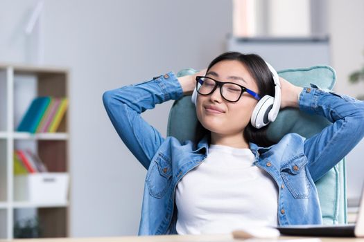 Young beautiful office worker relaxing sitting at the desk, Asian woman listening to music in headphones during work break.