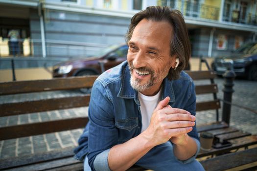 Handsome smiling mature man spend time talking on the phone or listening music using wireless earphones sitting on the bench in urban city, enjoying coffee wearing blue jeans shirt.