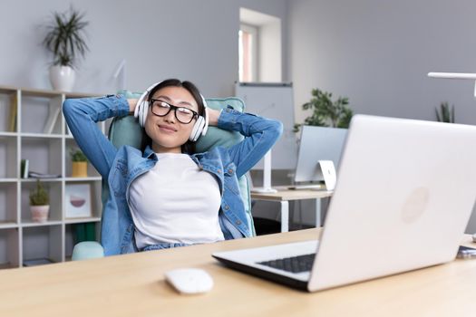 Young beautiful office worker relaxing sitting at the desk, Asian woman listening to music in headphones during work break.