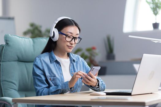 Young beautiful Asian woman listening to music in office, using phone and music app, businesswoman with headphones resting.